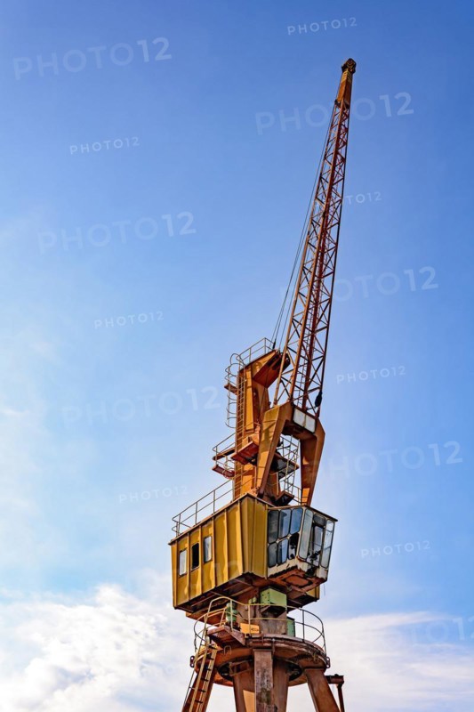 Old and obsolete yellow crane on the harbor pier with blue sky and clouds in the background.