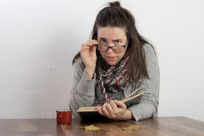 Young brunette woman with long hair and glasses reading a book holding her glasses and looking at the camera with a white background
