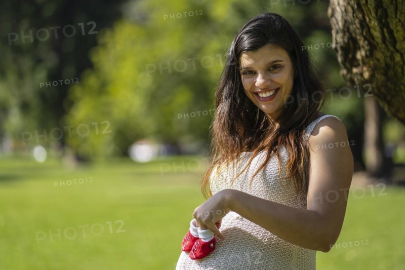 Pregnant latin woman holding baby shoes on her tummy in a park