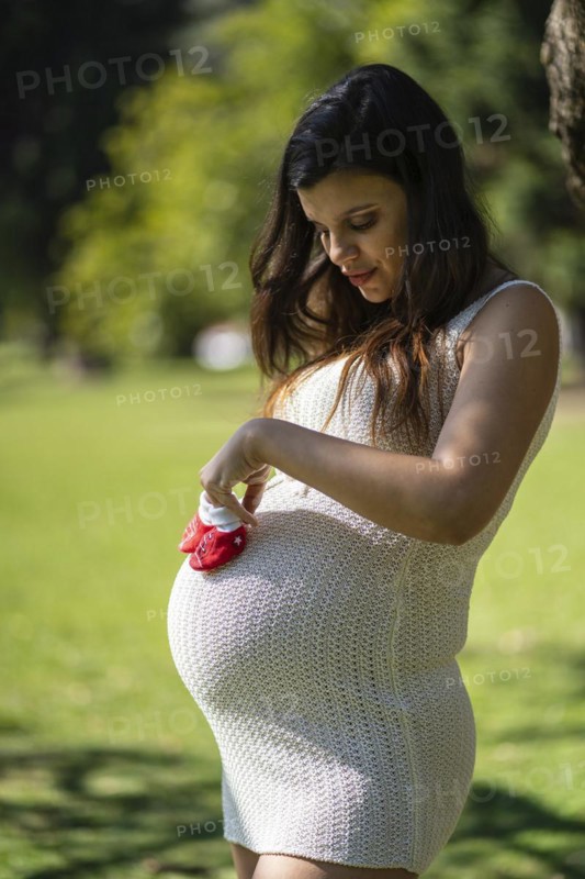 Pregnant latin woman holding baby shoes on her tummy in a park