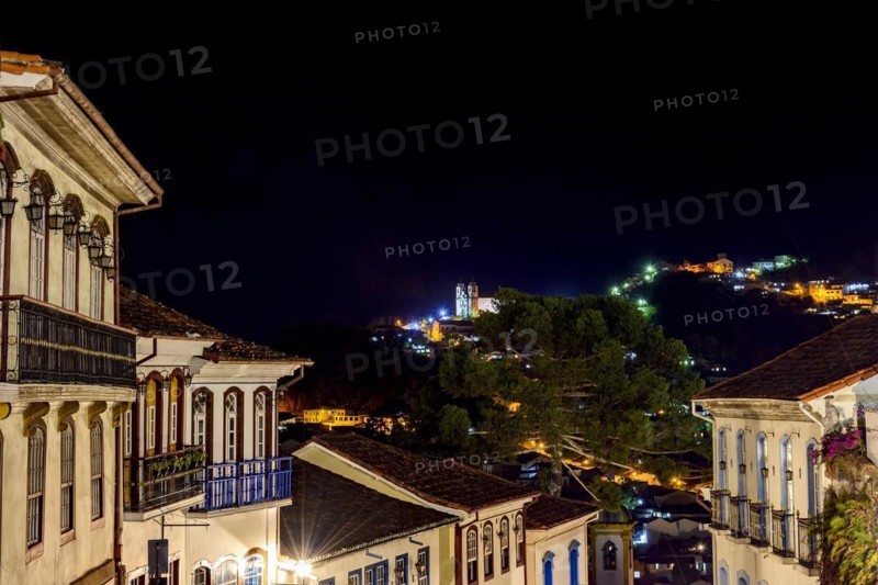 Facades of houses in colonial architecture on an old cobblestone street in the city of Ouro Preto illuminated at night with historic church in background