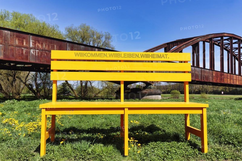 Oversized yellow wooden bench in front of old railway bridge