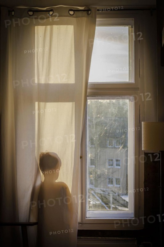 Symbolic photo on the subject of psychology in children. A boy stands at the window behind a curtain and looks out. Berlin
