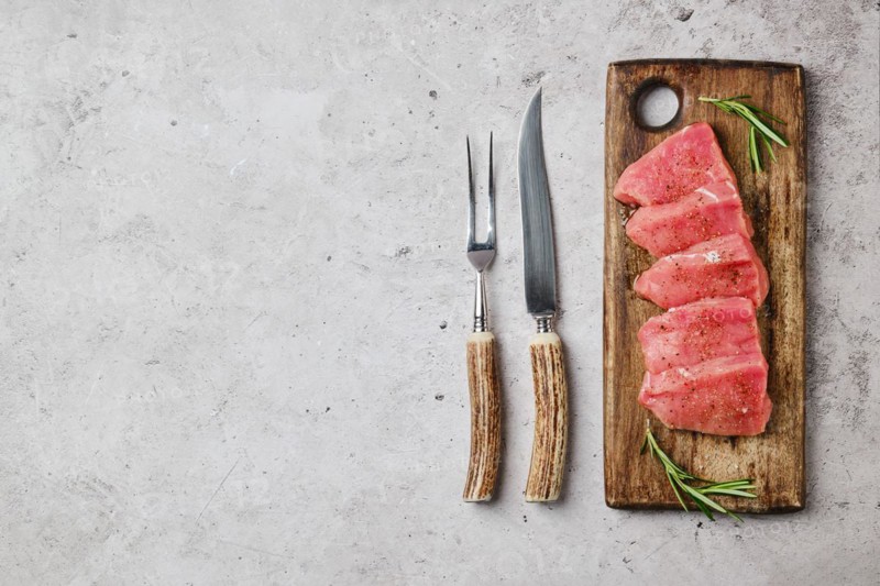 Top view of pork fillet sliced into pieces on wooden cutting board next to knife and fork for grill