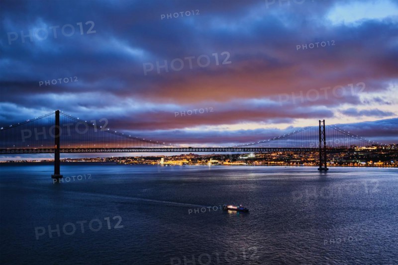 View of 25 de Abril Bridge famous tourist landmark of Lisbon connecting Lisboa and Almada on Setubal Peninsula over Tagus river in the evening twilight with tourist boats. Lisbon