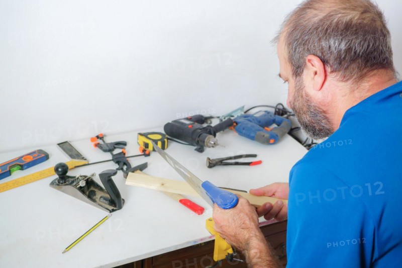Rear view of a male carpenter cutting wood in his workshop with a saw in the background with various tools on the table
