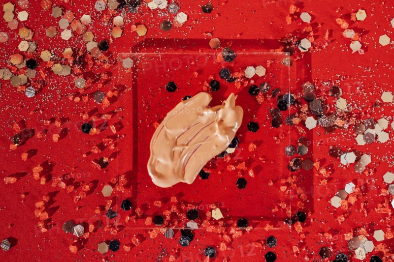 Cosmetic smear of tone cream on transperent glass. Bright red background with silver glitters and confetti. Concept of health and celebration. Copy space
