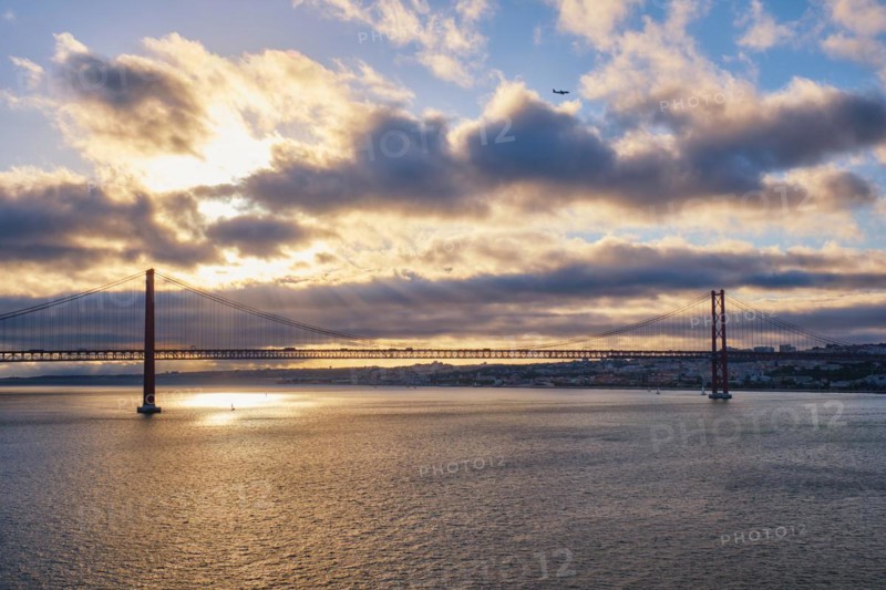 View of 25 de Abril Bridge famous tourist landmark of Lisbon connecting Lisboa and Almada on Setubal Peninsula over Tagus river on sunset with flying plane. Lisbon