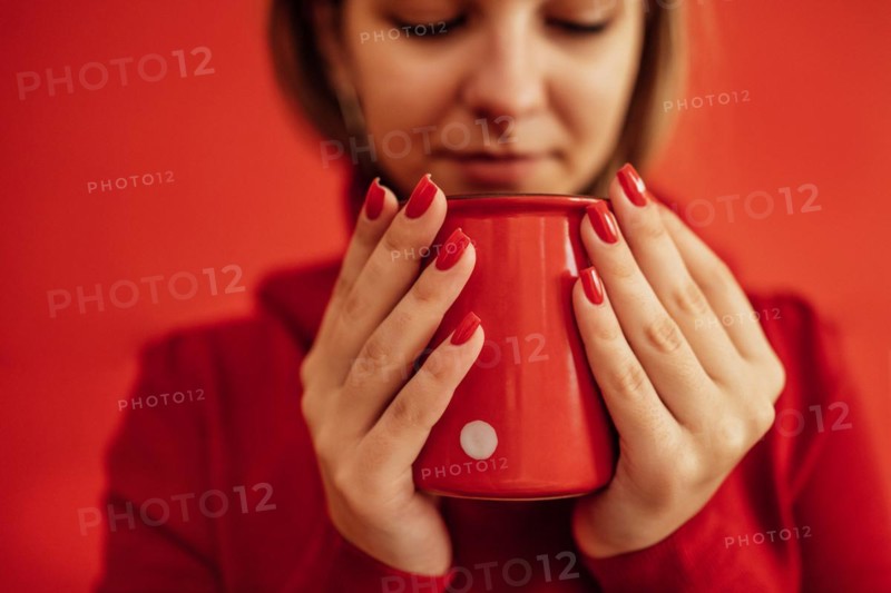 Young blondy girl in a red sweater holds a red cup in her hands. Close up of a beautiful bright red manicure. Valentine Day concept. Coffee break. Relax time