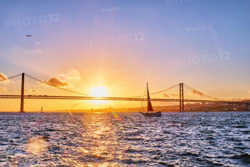View of 25 de Abril Bridge famous tourist landmark of Lisbon connects Lisboa and Almada on Setubal Peninsula over Tagus river with tourist yacht silhouette at sunset and flying plane. Lisbon