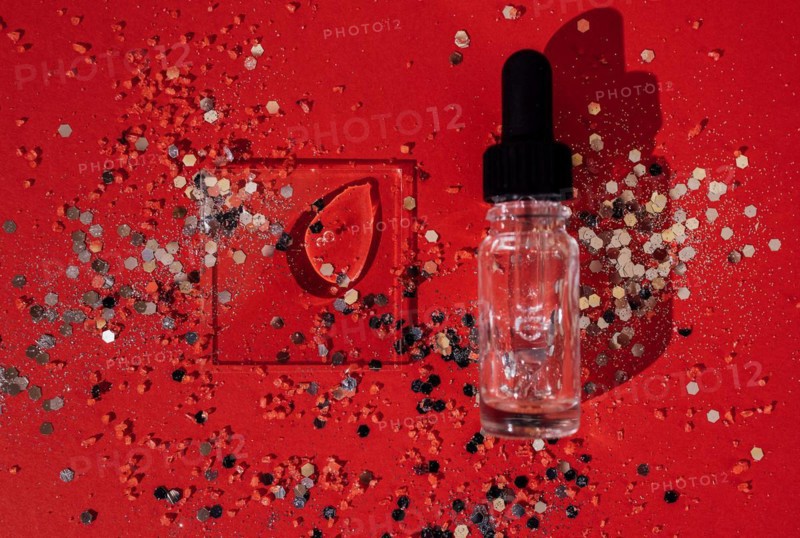 Transparent serum or oil glass bottle with pipette. Bright red background with silver glitters and confetti. Mock up. Concept of cosmetic and celebration. Copy space