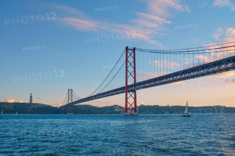 View of 25 de Abril Bridge famous tourist landmark of Lisbon connecting Lisboa to Almada on Setubal Peninsula over Tagus river with tourist yacht boats passing under. Lisbon