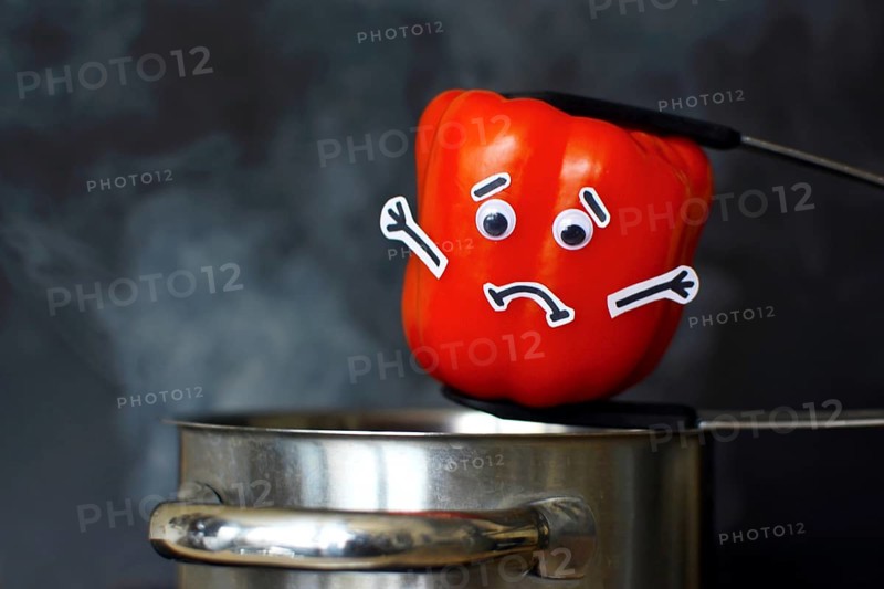 Red Paprika bell pepper with sad face and goggle eyes being put into a steaming cooking pot on dark black background