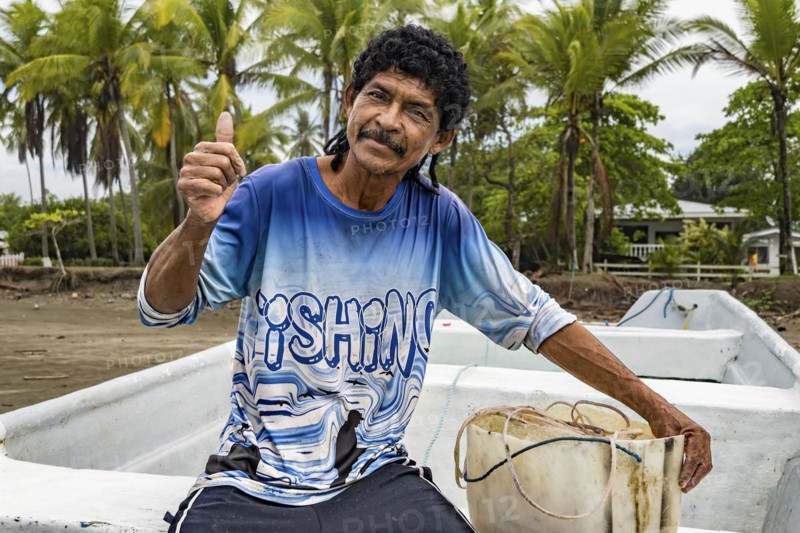 Man looking at camera with thumb up on his boat and fishing net. Background of palm trees out of focus