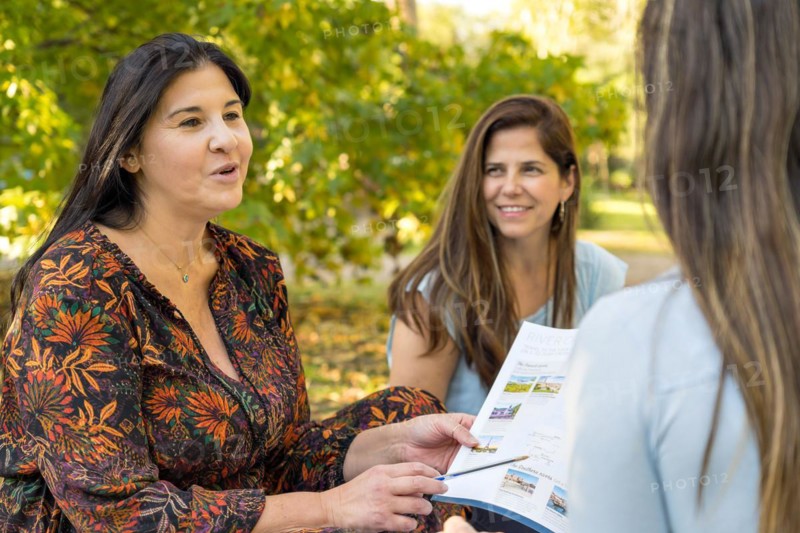 A female travel agent holds up an itinerary and explains vacation packages to two friends who listen attentively and excitedly