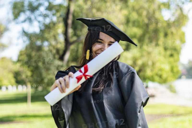 Happy caucasian graduated girl with long brown hair showing her diploma. She is wearing a bachelor gown and a black mortarboard