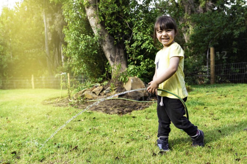 Portrait of happy smiling little child girl playing and watering garden grass with rubber strap and sunny summer in garden at home