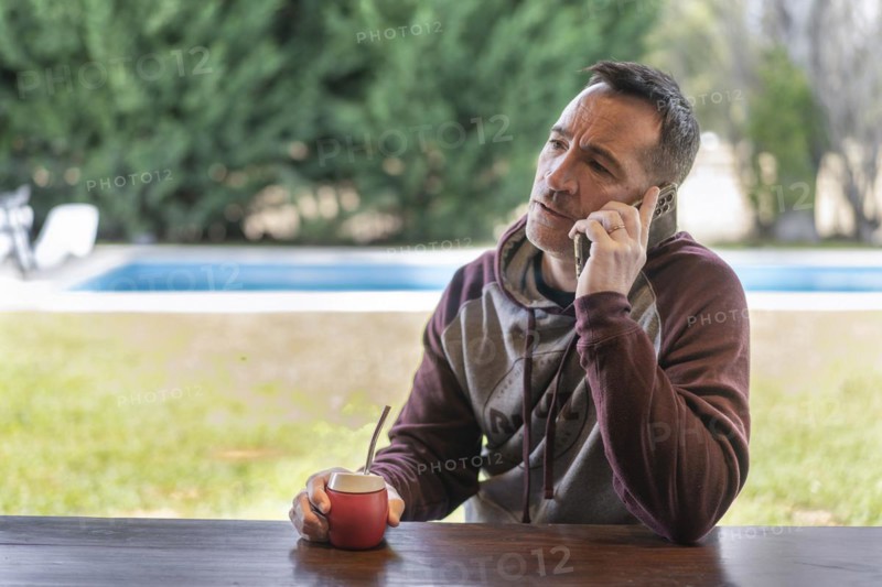 Serious adult man talking on the cellphone while drinking mate infusion at home