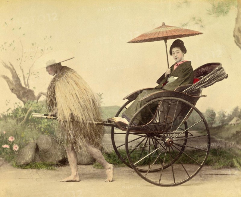 A distinguished woman with a parasol being pulled in a jinrikisha by a man wearing a straw mackintosh