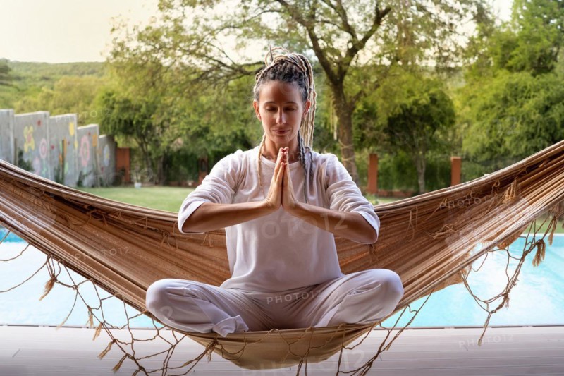 Blonde yogi woman with dreadlocks in prayer position sitting in a woven hammock