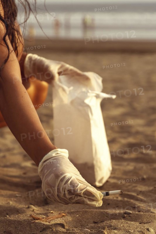 Volunteer girl collecting cigarette butts on the sand. Out-of-focus sunset background. Marine pollution destroying the environment