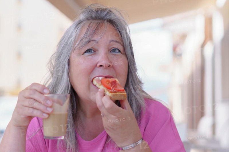 Close-up of an older white-haired woman drinking coffee with milk and eating a ham and tomato toast