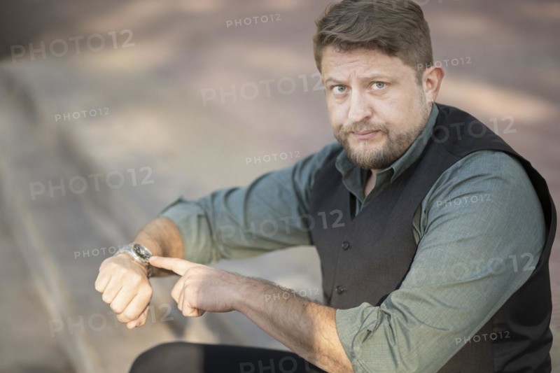 Man sitting in a park pointing at his watch looks disgusted at the camera. Delay concept