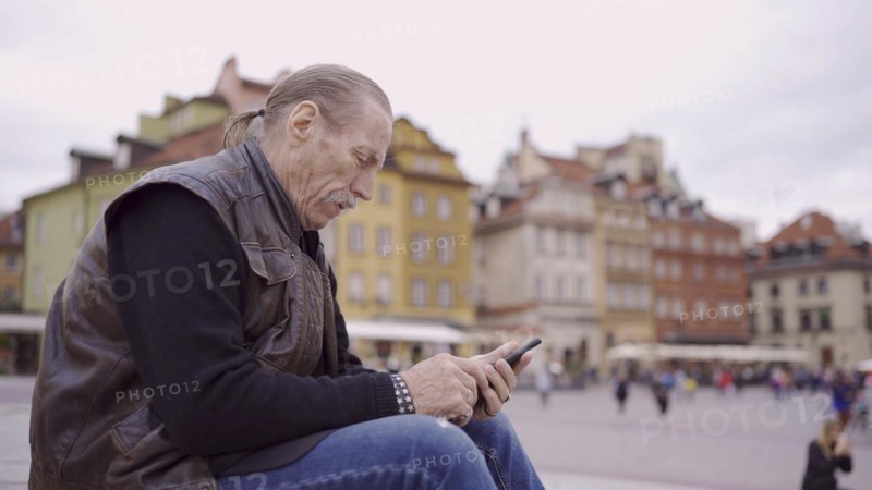 Senior sits on the steps and uses a smartphone in the historic center of an old European city