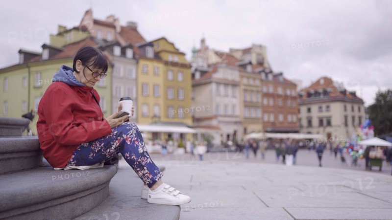 Elderly woman sits on the steps drinking coffee and using a smartphone in the historic center of an old European city