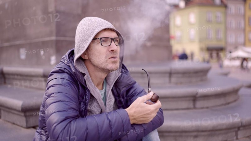 Adult man with glasses sitting on square and smoking a tobacco pipe releasing smoke in the Palace Square