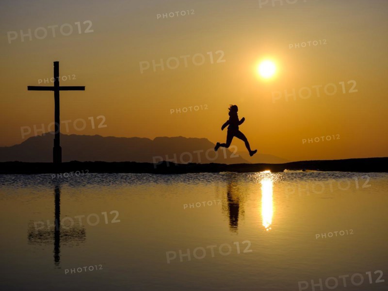 Silhouette of a woman jumping for joy next to a summit cross at sunset