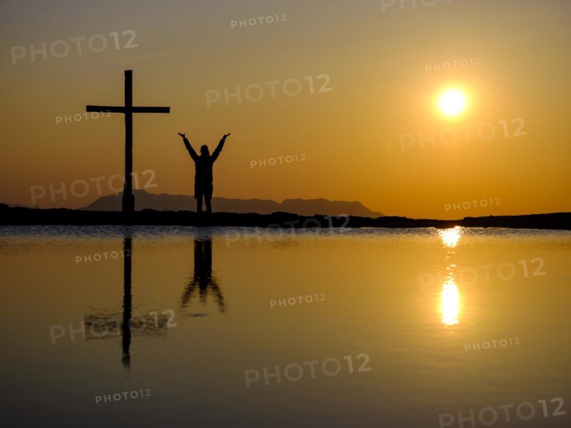 Silhouette of a woman with arms stretched up next to a cross on a summit at sunset