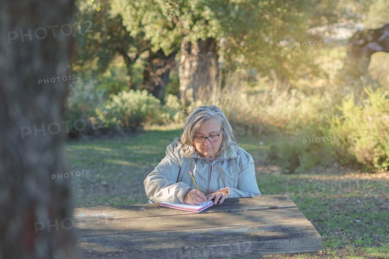 Front view of a white-haired woman sitting at a wooden table in a park writing in a notebook