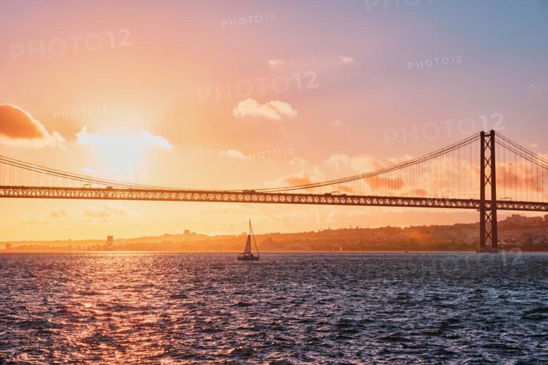View of 25 de Abril Bridge famous tourist landmark of Lisbon connecting Lisboa and Almada on Setubal Peninsula over Tagus river with tourist yacht silhouette at sunset