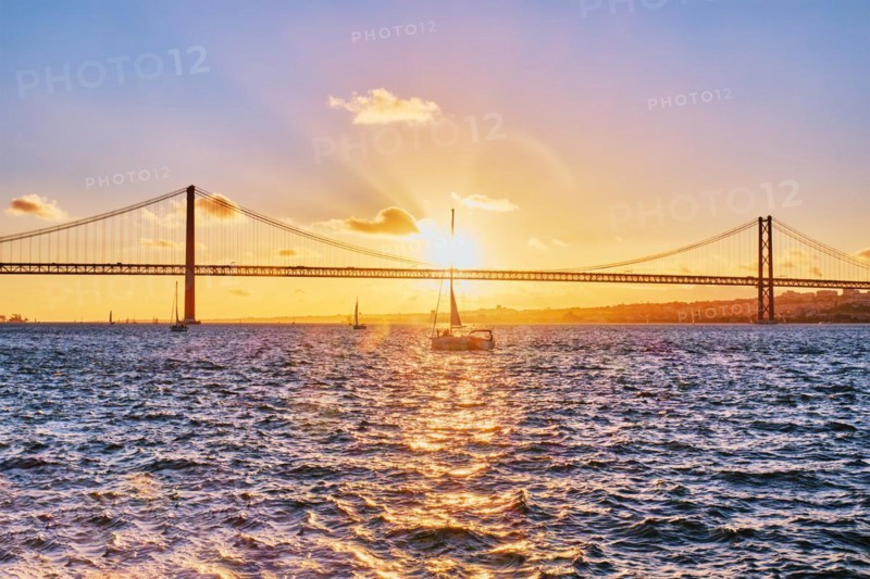 View of 25 de Abril Bridge famous tourist landmark of Lisbon connecting Lisboa and Almada on Setubal Peninsula over Tagus river with tourist yacht silhouette at sunset