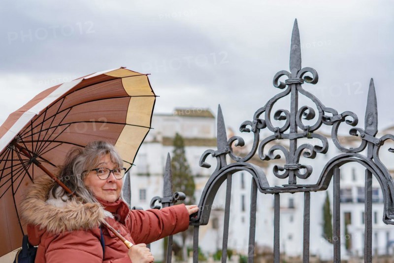 White-haired woman with coat and umbrella huddled against an iron fence on a dark and rainy autumn day