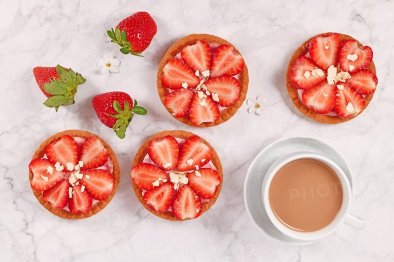 Small tartlet pastries topped with strawberry fruits and white chocolate sprinkles next to coffee cup