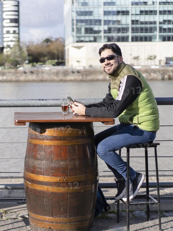 A smiling man looking at camera holding his cell phone at a bar near the river