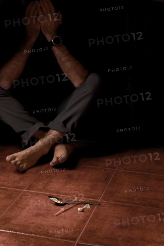 Unidentified barefoot man sitting on the floor in semi-darkness with a syringe and spoon containing heroin in the foreground