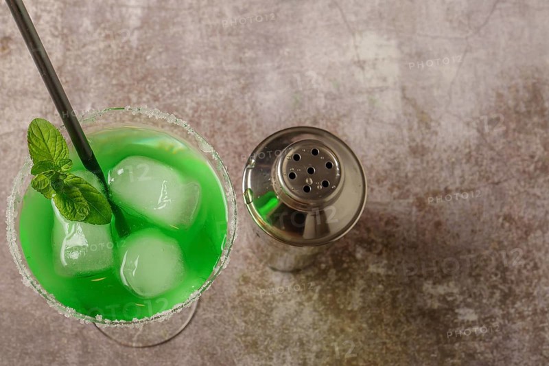 Top view of a green cocktail with mint ice and straw the edges of the glass have icing sugar and a cocktail shaker to accompany it