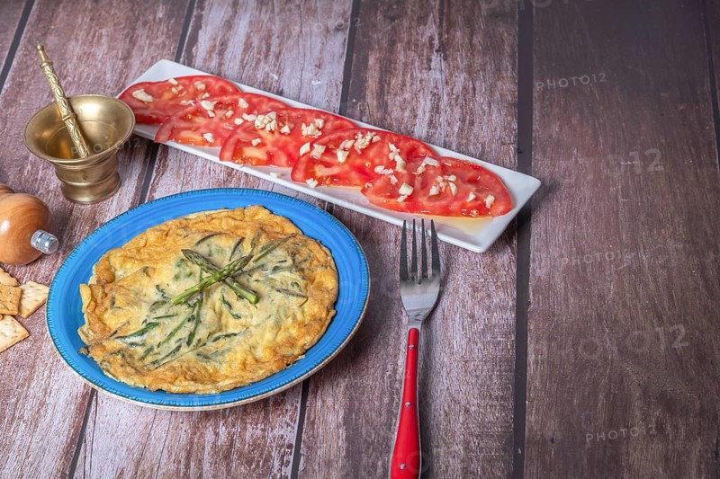 Wild asparagus omelette on a blue plate with a tomato salad with garlic on a wooden table
