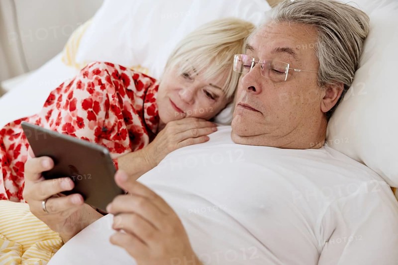 Elderly couple lying together in bed in bedroom looking at smartphone together