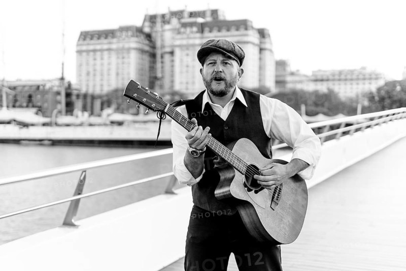 Black and white portrait of a musician playing the guitar and singing on a bridge over a river