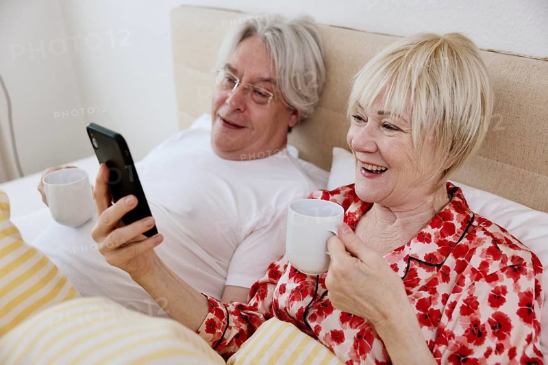 Elderly couple lying together in bed in bedroom looking at smartphone together