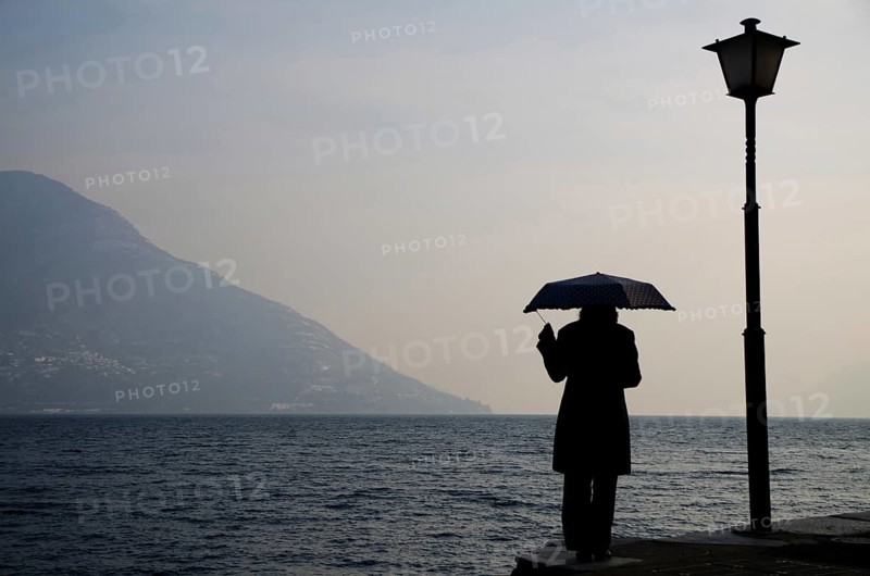 Woman Standing on the Waterfront with an Umbrella close to a Street Lamp