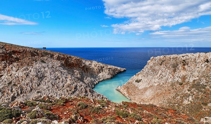 Beautiful view of z-shaped lagoon in typical hills of Crete landscape. Clear blue sky and beautiful clouds on sunny day. Azure sea waters. Red soil in foreground. Stefanou beach