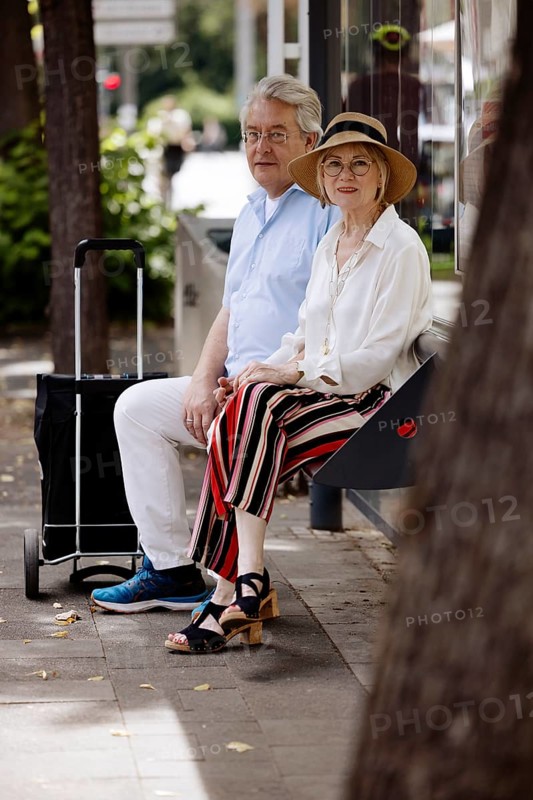 Older couple dressed in summer clothes with a trolley waiting for a bus at a bus stop