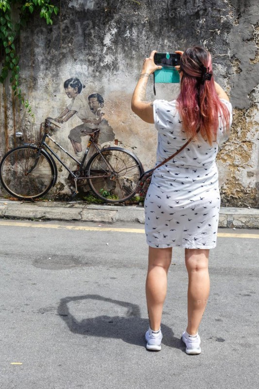 Young woman taking a photo of street art boy and girl on bicycle on a wall in George Town on Penang Island
