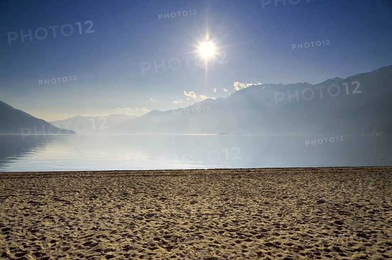 Sand Beach on a Misty Alpine Lake Maggiore with Mountain and Sunlight in Ascona