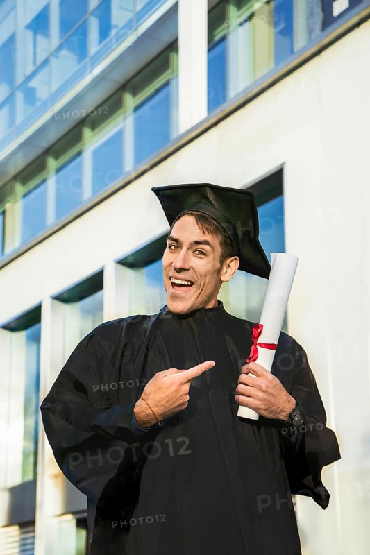 Happy graduated man wearing a bachelor gown and a black mortarboard and pointing at his diploma while looking at camera